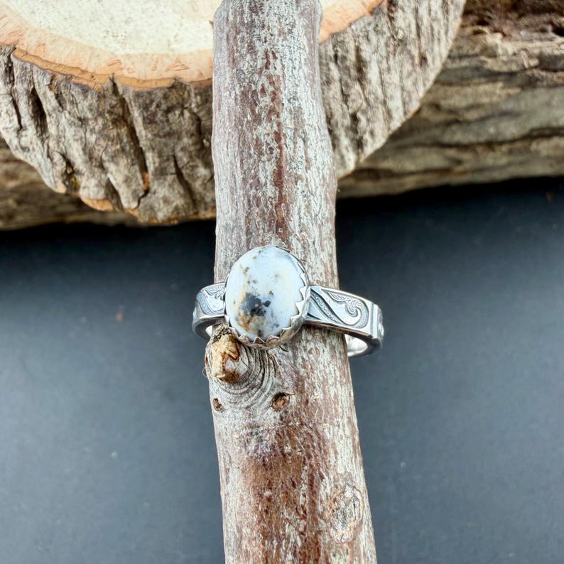 May include: A silver ring with an oval, light-colored stone featuring dark spots. The ring has a decorative band with etched patterns. The ring is displayed on a wooden branch against a dark background.