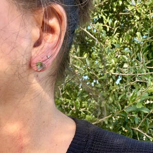 May include: A small, silver-toned heart-shaped stud earring with a textured, metallic surface. The earring is secured to the earlobe with a thin, curved metal wire that extends upward along the outer edge of the ear cartilage.