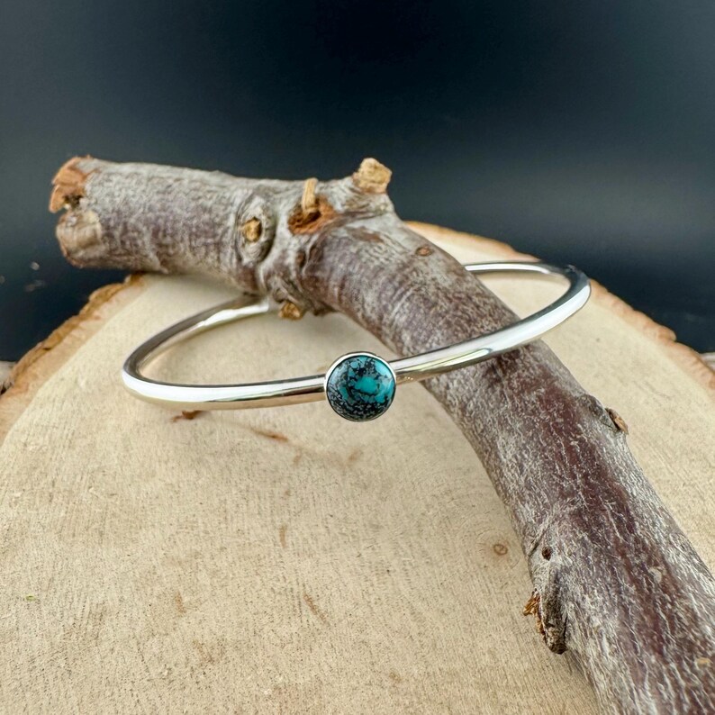 May include: A silver bangle bracelet with a round, turquoise-colored stone set in the center. The bracelet is displayed on a wooden surface with a dark background, highlighting the jewelry's design and color.