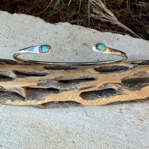 May include: A silver cuff bracelet featuring an open design with two polished, oval-shaped turquoise stones set at each end. The metal band has a smooth, reflective finish, and the turquoise stones display natural blue and brown veining.