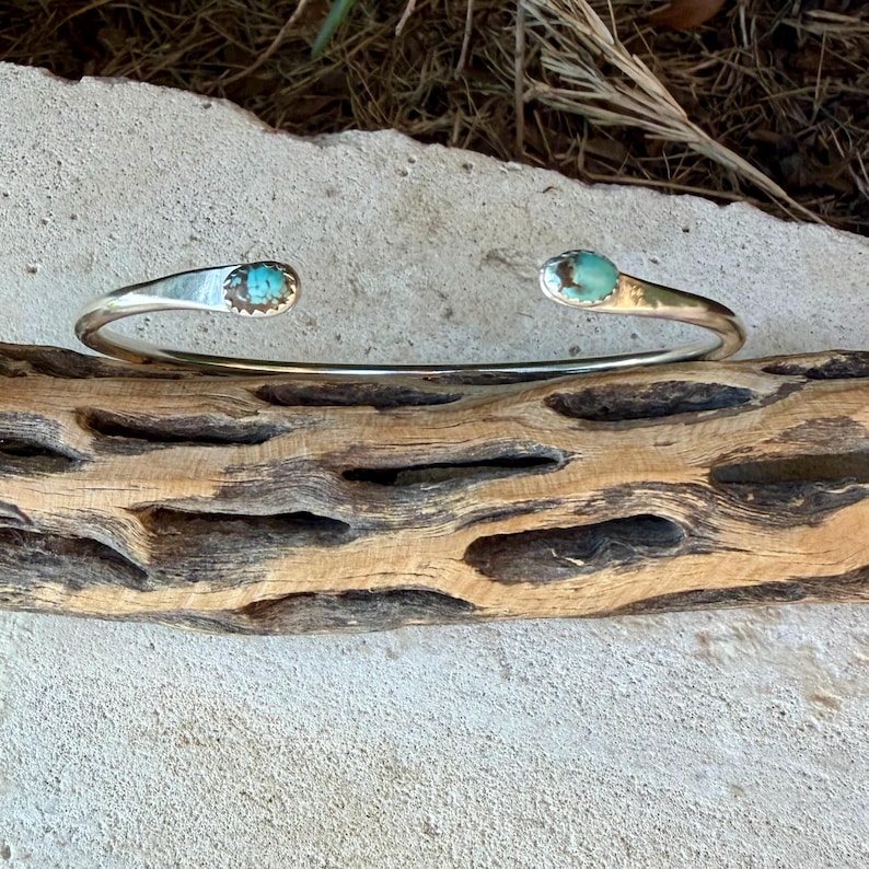 May include: A silver cuff bracelet featuring an open design with two polished, oval-shaped turquoise stones set at each end. The metal band has a smooth, reflective finish, and the turquoise stones display natural blue and brown veining.