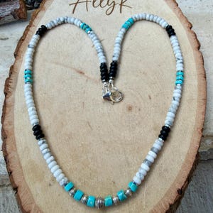 May include: A beaded necklace with white, black, and turquoise beads. The necklace has a silver clasp and is displayed on a wooden surface. The word "AllyK" is written in cursive at the top of the image.