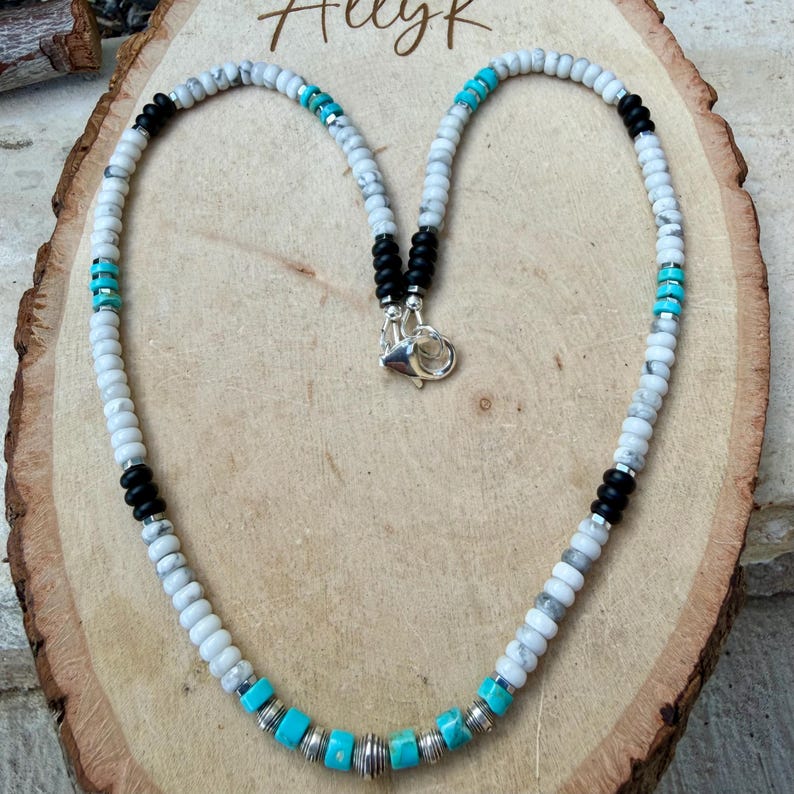 May include: A beaded necklace with white, black, and turquoise beads. The necklace has a silver clasp and is displayed on a wooden surface. The word "AllyK" is written in cursive at the top of the image.
