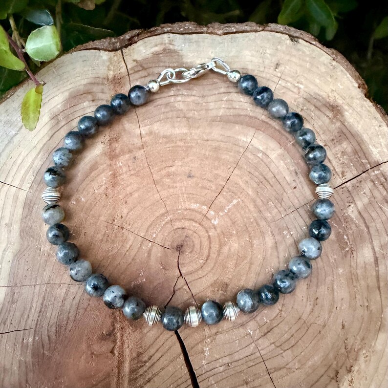 May include: A beaded bracelet featuring round, dark gray, mottled stone beads interspersed with small, ribbed, silver-toned metal spacer beads. The bracelet is secured with a silver-toned metal lobster claw clasp and a jump ring.