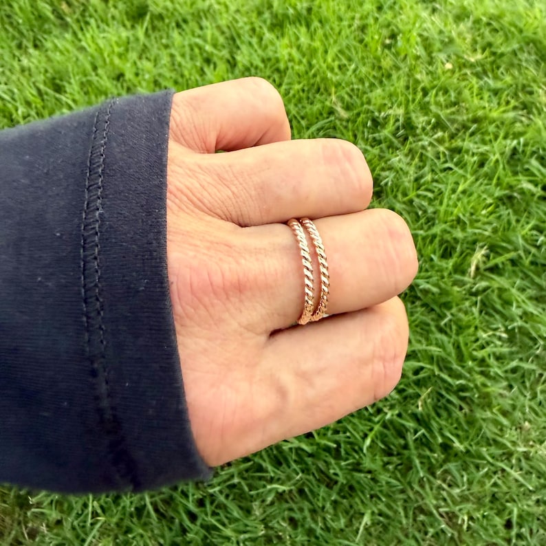 May include: Two gold-colored, twisted rope design rings are worn on a finger. The rings are made of a shiny metal and are stacked together. The background is blurred green grass.