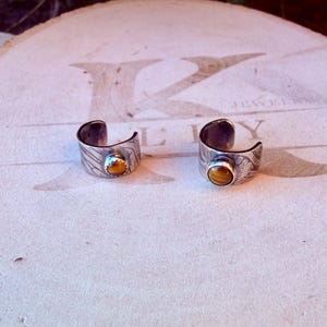 K&ouml;nnte beinhalten: Zwei offene Ringe aus silberfarbenem Metall mit einer strukturierten, wellenf&ouml;rmigen Oberfl&auml;che. Jeder Ring ist mit einem einzelnen, runden, polierten Tigerauge-Edelstein besetzt, der in einer gezackten Fassung sitzt.