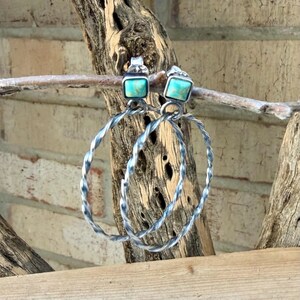 May include: A pair of silver hoop earrings with square turquoise stones. The hoops have a twisted design. The earrings are displayed on a wooden branch against a brick wall background.