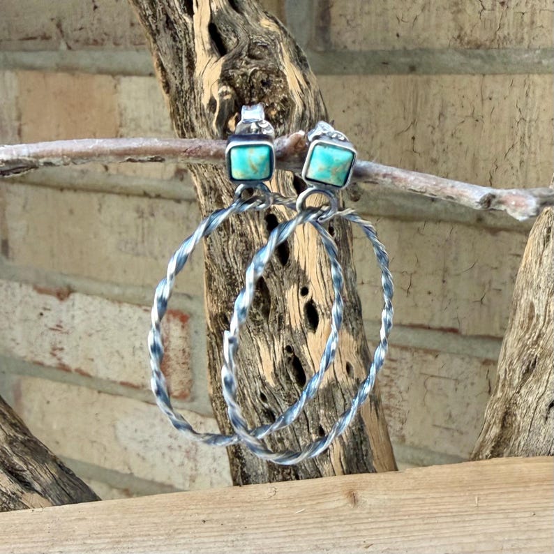 May include: A pair of silver hoop earrings with square turquoise stones. The hoops have a twisted design. The earrings are displayed on a wooden branch against a brick wall background.