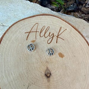 Puede incluir: Un par de pendientes de bot&oacute;n plateados con el s&iacute;mbolo de la paz. Los pendientes se muestran sobre una superficie de madera con el texto escrito a mano "Allyk". Los pendientes son peque&ntilde;os y redondos, con un s&iacute;mbolo de la paz en el centro.
