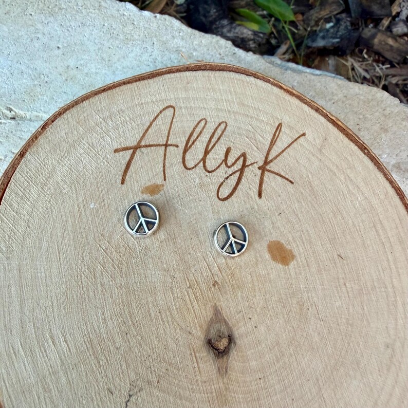 Puede incluir: Un par de pendientes de bot&oacute;n plateados con el s&iacute;mbolo de la paz. Los pendientes se muestran sobre una superficie de madera con el texto escrito a mano "Allyk". Los pendientes son peque&ntilde;os y redondos, con un s&iacute;mbolo de la paz en el centro.