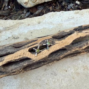 K&ouml;nnte beinhalten: Ein Paar silberfarbene Ohrstecker in Kreuzform mit einem kleinen, runden, hellgr&uuml;nen Stein in der Mitte. Die Ohrringe werden auf einem St&uuml;ck verwittertem, hellbraunem Holz pr&auml;sentiert.