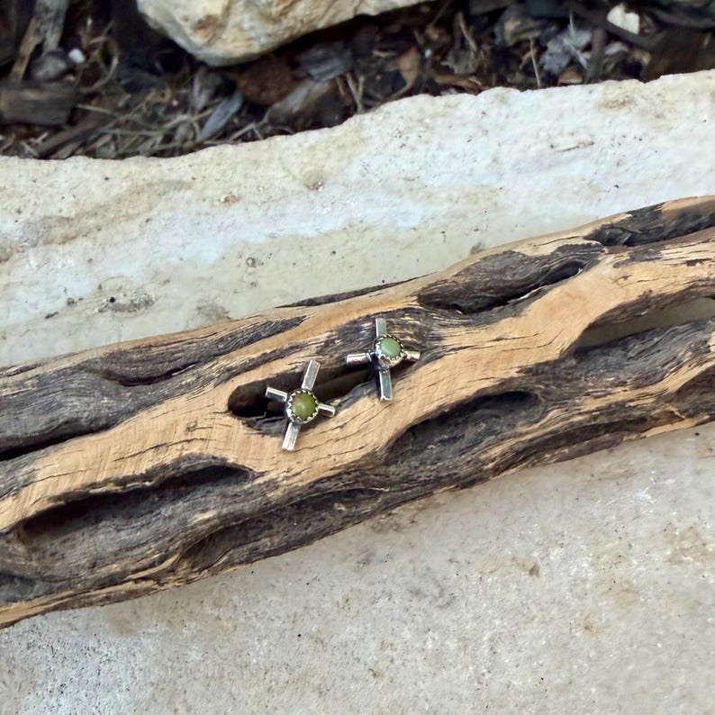 K&ouml;nnte beinhalten: Ein Paar silberfarbene Ohrstecker in Kreuzform mit einem kleinen, runden, hellgr&uuml;nen Stein in der Mitte. Die Ohrringe werden auf einem St&uuml;ck verwittertem, hellbraunem Holz pr&auml;sentiert.