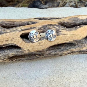 May include: A pair of round silver stud earrings, each featuring a stamped sun symbol design with a central dot and four radiating lines. The earrings have a polished metallic finish and are circular in shape, displaying a clear, symmetrical pattern on the front surface.