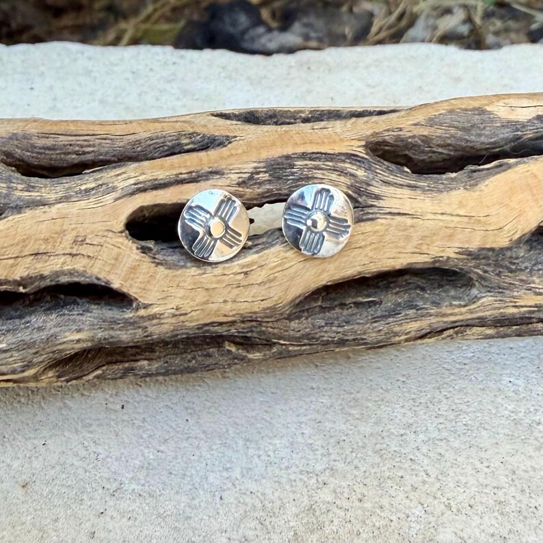 May include: A pair of round silver stud earrings, each featuring a stamped sun symbol design with a central dot and four radiating lines. The earrings have a polished metallic finish and are circular in shape, displaying a clear, symmetrical pattern on the front surface.