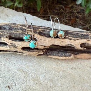 May include: Two pairs of drop earrings featuring round turquoise stones with brown matrix patterns set in silver-toned metal. Each earring has a curved wire hook for attachment, showcasing a polished finish on the stones and a simple metal setting.