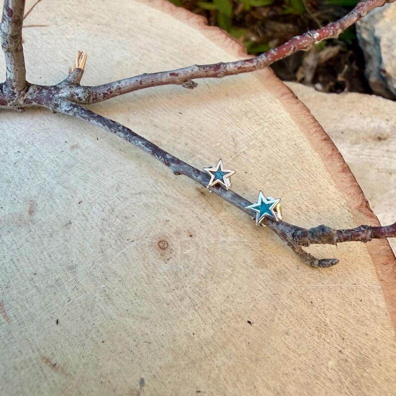 May include: A pair of star-shaped stud earrings with a silver-colored outline and a turquoise center. The earrings are displayed on a small tree branch against a light brown wooden background.