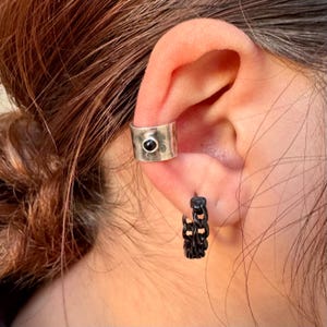 May include: A silver ear cuff with a black stone and a black chain hoop earring are shown on an ear. The ear cuff is wide and has a hammered texture. The hoop earring is made of a chain link design.