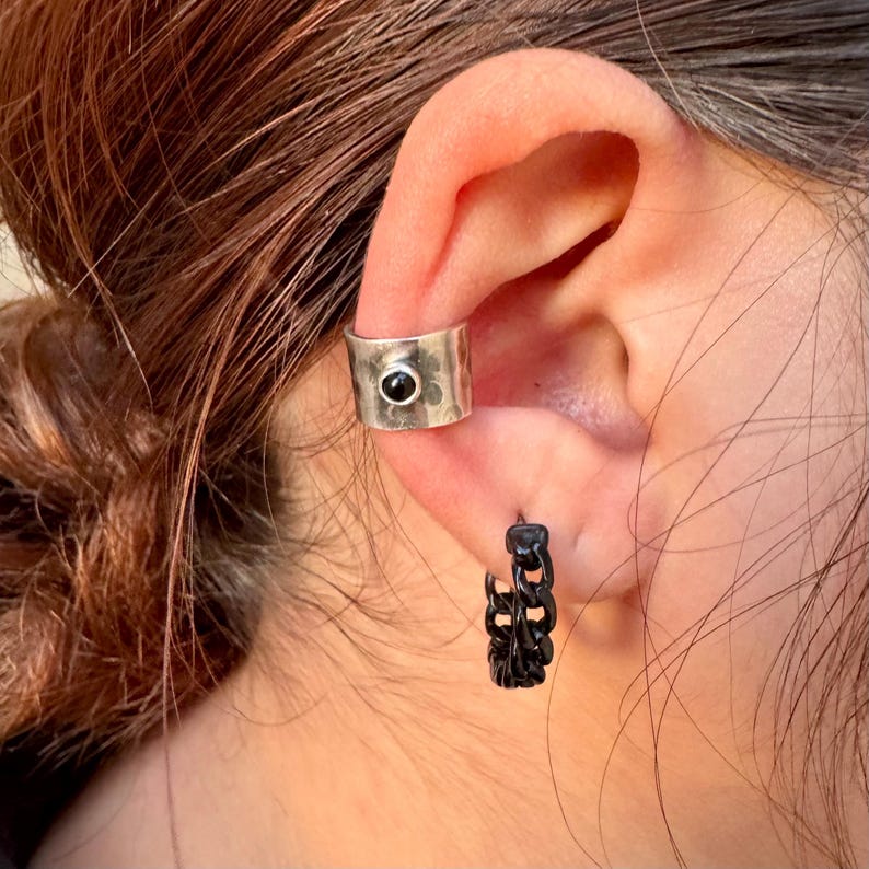 May include: A silver ear cuff with a black stone and a black chain hoop earring are shown on an ear. The ear cuff is wide and has a hammered texture. The hoop earring is made of a chain link design.