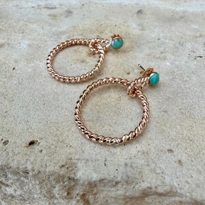 May include: A pair of rose gold hoop earrings with a twisted rope design. Each earring features a small, round turquoise stone at the top. The earrings are displayed on a textured, light-colored surface.