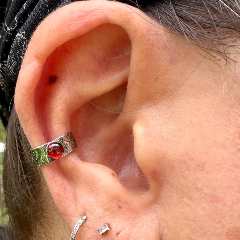 May include: A silver ear cuff with a round, red gemstone is worn on the ear. The cuff has a textured design. A small, silver hoop earring and a small, rectangular stud earring are also visible.