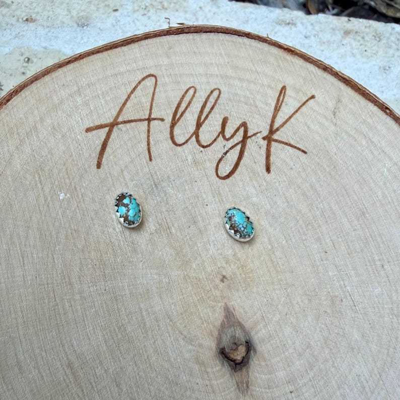 May include: A pair of oval turquoise stud earrings with a silver-colored border, displayed on a wooden surface. The earrings have a textured appearance, and the name "Allyk" is written in cursive on the wood.