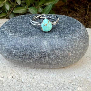 Puede incluir: Un anillo de plata con una banda de metal trenzado y una piedra turquesa &uacute;nica en forma de l&aacute;grima. La piedra es de color azul claro con vetas de matriz marr&oacute;n distintivas en toda su superficie. La banda tiene un acabado met&aacute;lico pulido.