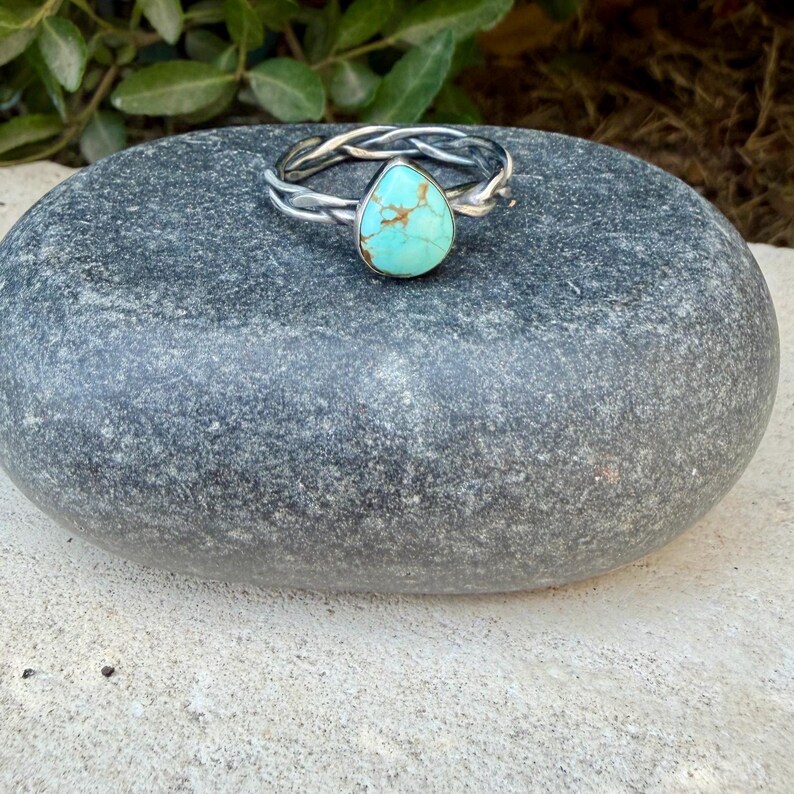 Puede incluir: Un anillo de plata con una banda de metal trenzado y una piedra turquesa &uacute;nica en forma de l&aacute;grima. La piedra es de color azul claro con vetas de matriz marr&oacute;n distintivas en toda su superficie. La banda tiene un acabado met&aacute;lico pulido.