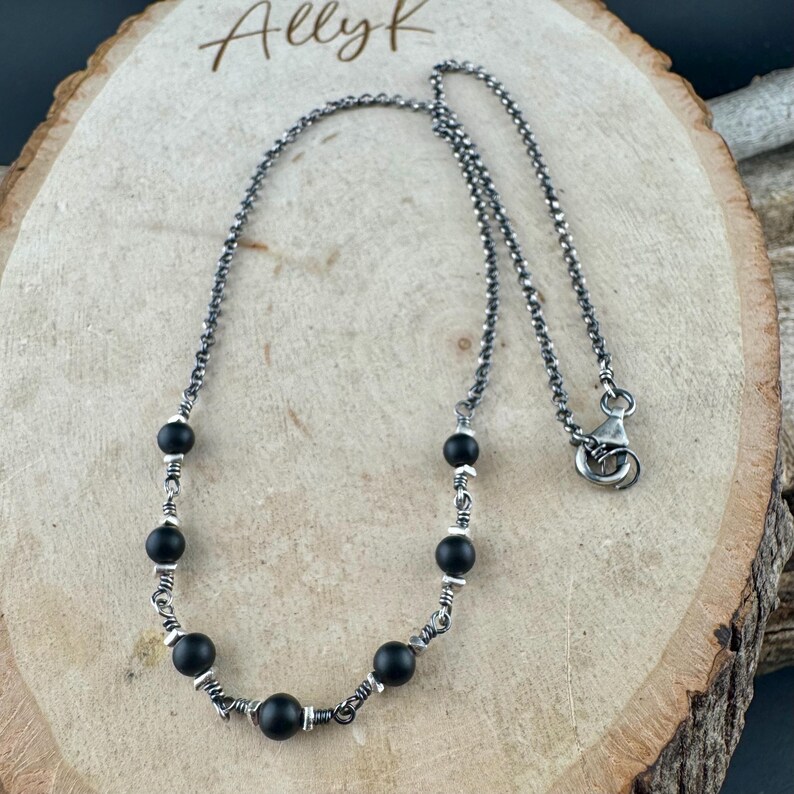 May include: A silver-toned necklace featuring black beads and a delicate chain. The necklace has a lobster clasp closure and is displayed on a wooden surface. The text "Ally K" is visible in the upper left corner.