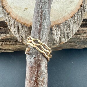 May include: A gold-colored ring with a wavy design is displayed on a wooden branch. The ring has a unique, flowing pattern, and the wood has a natural, textured appearance. The background is a dark color.