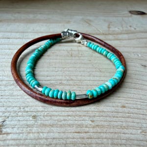 May include: A double-strand bracelet featuring one strand of smooth brown leather cord and one strand of small, round, turquoise-colored beads with subtle brown veining, accented by silver-toned metal spacers and a silver-toned lobster claw clasp.