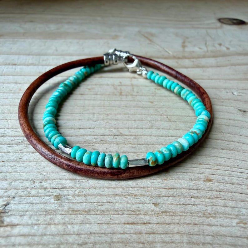 May include: A double-strand bracelet featuring one strand of smooth brown leather cord and one strand of small, round, turquoise-colored beads with subtle brown veining, accented by silver-toned metal spacers and a silver-toned lobster claw clasp.