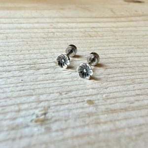 May include: A pair of silver-toned stud earrings featuring a small, multi-petaled flower design. Each earring has a central post and a rounded backing, with oxidized detailing within the grooves of the petals to define the floral shape.