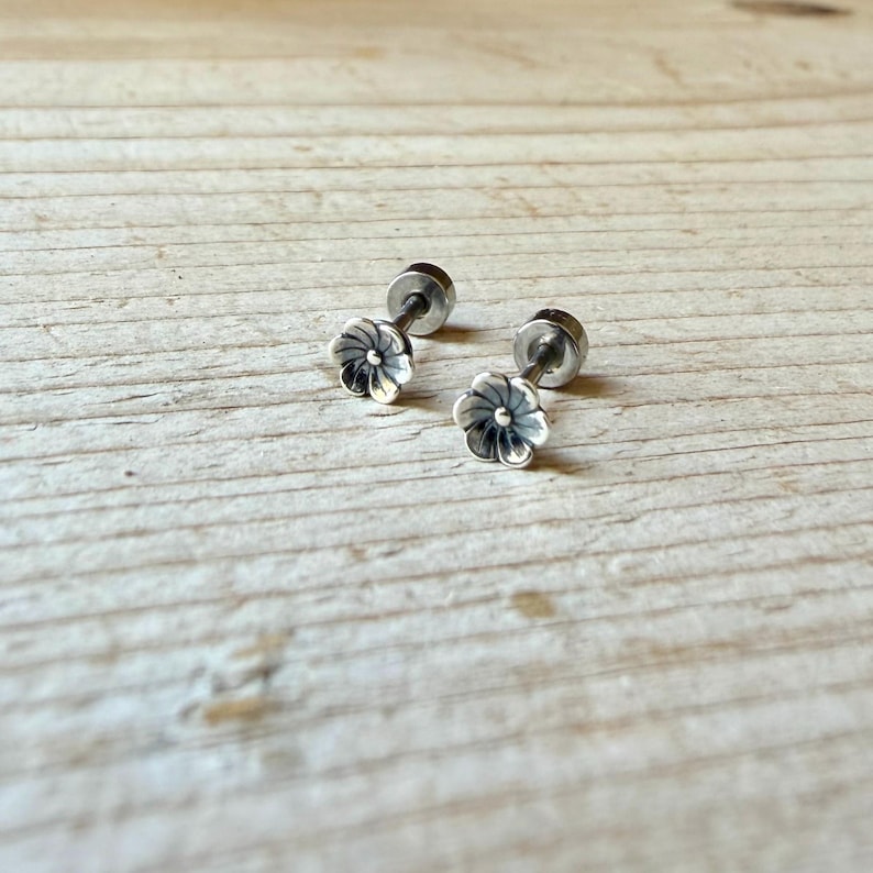 May include: A pair of silver-toned stud earrings featuring a small, multi-petaled flower design. Each earring has a central post and a rounded backing, with oxidized detailing within the grooves of the petals to define the floral shape.