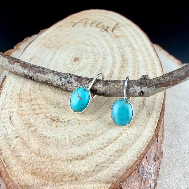 May include: A pair of silver-tone earrings with oval turquoise stones. The earrings are displayed on a wooden surface with a small branch. The word "Allyk" is written on the wood.
