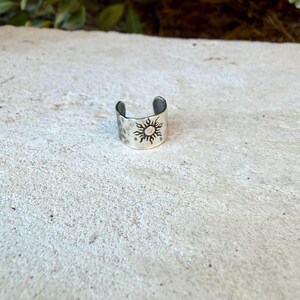May include: A silver-toned metal ring with an open band design, featuring a stamped sun motif with radiating rays on the front surface. The metal has a slightly textured, hammered finish, and the band is open at the back for adjustable sizing.
