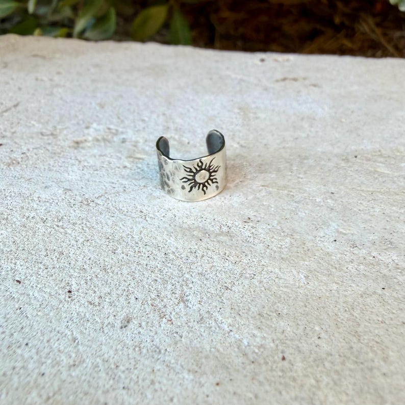 May include: A silver-toned metal ring with an open band design, featuring a stamped sun motif with radiating rays on the front surface. The metal has a slightly textured, hammered finish, and the band is open at the back for adjustable sizing.
