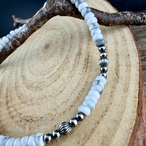 May include: A white and gray beaded necklace with silver-colored accents. The necklace features round and cylindrical beads, with a mix of matte and metallic finishes. The necklace is displayed on a wooden surface.