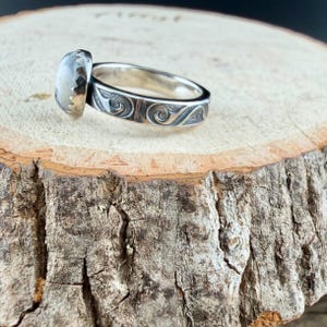 May include: A silver ring with an oval, light-colored stone. The band features an engraved, swirling pattern. The ring is set on a wooden surface, showcasing the jewelry's design and craftsmanship.