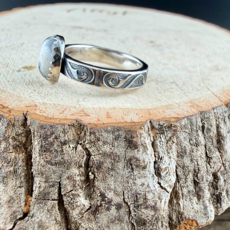May include: A silver ring with an oval, light-colored stone. The band features an engraved, swirling pattern. The ring is set on a wooden surface, showcasing the jewelry's design and craftsmanship.