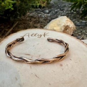 May include: A copper-colored, braided cuff bracelet. The open-ended bracelet is made of twisted metal strands. The bracelet sits on a wooden surface with the name "Ally K" engraved on it. The background is blurred with greenery and a rock.