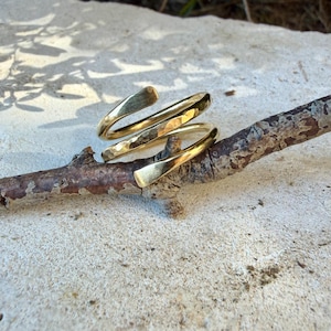 May include: A gold-toned metal ring with a bypass spiral design. The band features a hammered, textured finish on the outer surface, creating a reflective appearance. The ring is open-ended, with two tapered ends that overlap slightly in a coil shape.