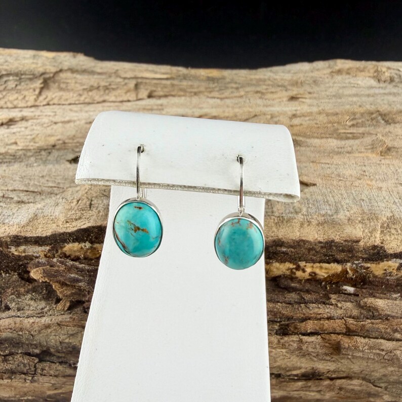 May include: A pair of silver drop earrings with round, turquoise-colored stones. The earrings are displayed on a white stand against a backdrop of wood.
