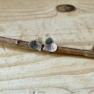 May include: A pair of silver heart-shaped stud earrings featuring an intricate, engraved swirling pattern on the surface. The earrings are positioned side-by-side, resting on a thin, textured brown twig.