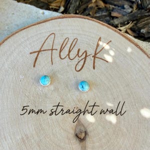 May include: A pair of turquoise stud earrings with a silver bezel, displayed on a wooden surface. The earrings are 5mm in size and are labeled "Allyk" and "5mm straight wall" in brown script.