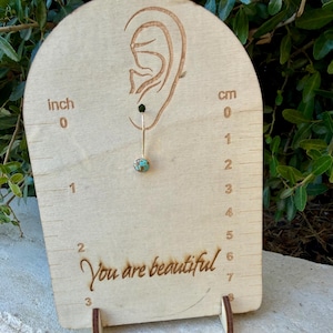 May include: A single dangle earring featuring a small, round turquoise stone with brown matrix set in a gold-toned metal bezel. The earring hangs from a thin gold-toned wire on a light wood display card with an ear outline and the text You are beautiful.