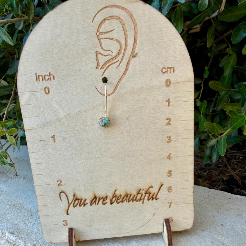May include: A single dangle earring featuring a small, round turquoise stone with brown matrix set in a gold-toned metal bezel. The earring hangs from a thin gold-toned wire on a light wood display card with an ear outline and the text You are beautiful.