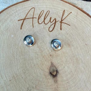 May include: A pair of silver-coloured cowboy hat stud earrings. The earrings are presented on a light brown wooden surface with the handwritten text "Allyk" in brown cursive script.