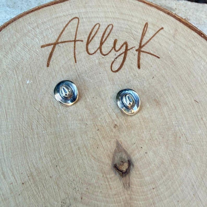May include: A pair of silver-coloured cowboy hat stud earrings. The earrings are presented on a light brown wooden surface with the handwritten text "Allyk" in brown cursive script.