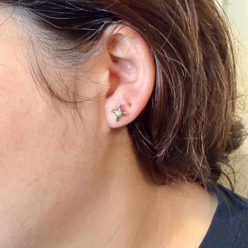K&ouml;nnte beinhalten: Nahaufnahme eines silbernen Ohrsteckers in Kreuzform mit einem kleinen, hellgr&uuml;nen Stein in der Mitte. Der Ohrring wird am Ohr einer Person mit dunkelbraunem Haar getragen.