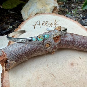 May include: A silver cuff bracelet with a hammered texture and four turquoise stones. The bracelet is displayed on a wooden surface with the handwritten text "Ally K". The turquoise stones are a mix of blue and brown.